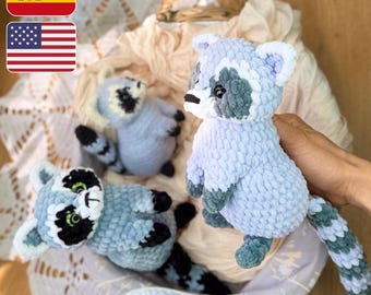 May include: Three handmade crochet raccoon plush toys in shades of blue, gray, and white. The raccoons have black and white facial markings, and long, striped tails. The toys are displayed in a basket with a cream-colored lining.