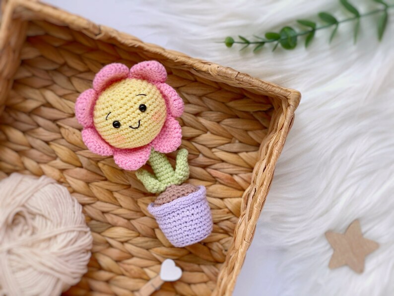 May include: A crocheted flower with a smiling face, pink petals, yellow center, and green leaves. The flower is in a purple pot.