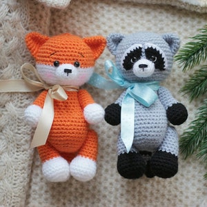Puede incluir: Dos animales de peluche tejidos a mano, un zorro y un mapache. El zorro es naranja y blanco con una cinta beige. El mapache es gris, negro y blanco con una cinta azul claro de lunares. Ambos tienen ojos de pl&aacute;stico negro y narices bordadas.