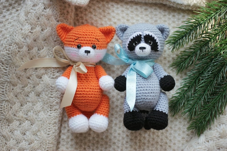 Puede incluir: Dos animales de peluche tejidos a mano, un zorro y un mapache. El zorro es naranja y blanco con una cinta beige. El mapache es gris, negro y blanco con una cinta azul claro de lunares. Ambos tienen ojos de pl&aacute;stico negro y narices bordadas.