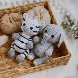 May include: Two handmade crochet stuffed animals, a zebra and an elephant, sitting together. The zebra has black and white stripes with gray accents, while the elephant is solid light gray. Both have black safety eyes and pink stitched cheeks.