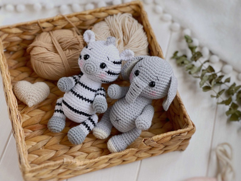 May include: Two handmade crochet stuffed animals, a zebra and an elephant, sitting together. The zebra has black and white stripes with gray accents, while the elephant is solid light gray. Both have black safety eyes and pink stitched cheeks.