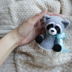 Puede incluir: Un peque&ntilde;o peluche de mapache tejido a ganchillo en color gris con parches negros en los ojos, patas negras y hocico blanco. El mapache lleva una cinta azul claro alrededor del cuello y est&aacute; dentro del bolsillo de un su&eacute;ter de punto crema. Texto: @KNIT.FRIENDS.