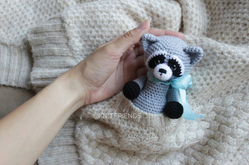 Puede incluir: Un peque&ntilde;o peluche de mapache tejido a ganchillo en color gris con parches negros en los ojos, patas negras y hocico blanco. El mapache lleva una cinta azul claro alrededor del cuello y est&aacute; dentro del bolsillo de un su&eacute;ter de punto crema. Texto: @KNIT.FRIENDS.