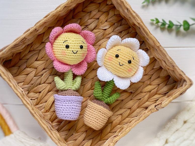 May include: Two crocheted flower pot decorations. One flower is pink with a yellow center and the other is white with a yellow center. Both flowers have smiling faces and are in small pots.