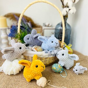 Puede incluir: Cinco conejos de crochet, cuatro grises y uno amarillo, est&aacute;n dispuestos alrededor de una cesta de mimbre con huevos de Pascua. El conejo amarillo sostiene una bola de hilo.