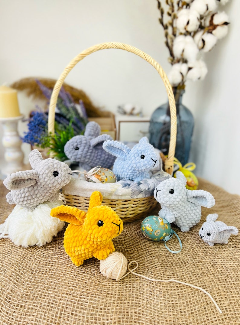Puede incluir: Cinco conejos de crochet, cuatro grises y uno amarillo, est&aacute;n dispuestos alrededor de una cesta de mimbre con huevos de Pascua. El conejo amarillo sostiene una bola de hilo.
