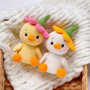 May include: Two small crocheted ducklings, one yellow and one white, each wearing a green and pink or yellow flower-shaped hat. Both figures have orange beaks and feet, black eyes, and pink blush on their cheeks, crafted with visible yarn stitches.