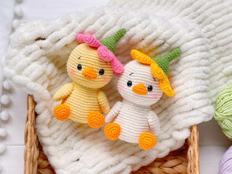 May include: Two small crocheted ducklings, one yellow and one white, each wearing a green and pink or yellow flower-shaped hat. Both figures have orange beaks and feet, black eyes, and pink blush on their cheeks, crafted with visible yarn stitches.
