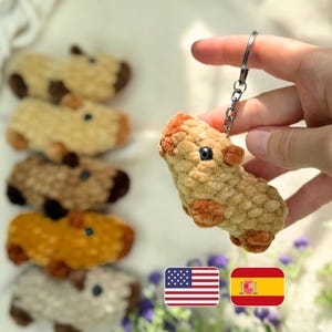May include: A handmade, plush capybara keychain in shades of tan and brown, held by a hand. The keychain features a silver ring and chain. Several other capybara plushies in various colors are visible in the background.