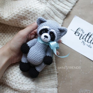 Puede incluir: Un juguete de mapache tejido a ganchillo en gris y negro con una cinta azul claro atada al cuello. Junto al juguete hay una tarjeta blanca con el texto My knitting My rules y el nombre @KNIT.FRIENDS impresos en tinta negra sobre la tarjeta.