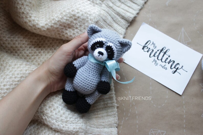 Puede incluir: Un juguete de mapache tejido a ganchillo en gris y negro con una cinta azul claro atada al cuello. Junto al juguete hay una tarjeta blanca con el texto My knitting My rules y el nombre @KNIT.FRIENDS impresos en tinta negra sobre la tarjeta.