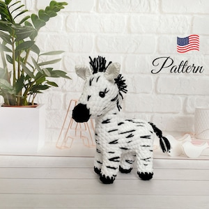 May include: A crocheted zebra toy in white with black stripes, mane, and tail. The word "Pattern" is written in cursive. The background features a white brick wall and a potted plant. The toy is a soft, handmade item.