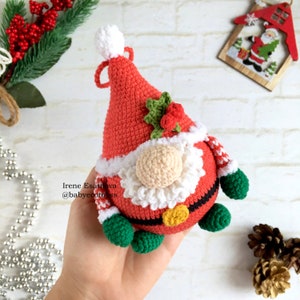 Pu&ograve; includere: Ornamento natalizio a forma di gnomo all'uncinetto. Lo gnomo ha un cappello rosso con un pompon bianco e una barba bianca. Ha un naso beige, piedi verdi e una cintura nera con una fibbia dorata. Lo gnomo tiene un rametto di agrifoglio.