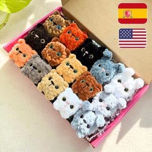 May include: A collection of handmade, plush cat-shaped toys in a variety of colors, including orange, black, and gray. The toys are arranged in a pink-lined cardboard box. Each cat has embroidered facial features.
