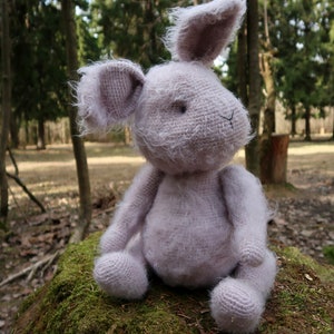 Op de afbeelding: Een zacht, pluche, lichtpaarse gehaakte konijnenknuffel die op een mossige boomstronk in een bosrijke omgeving zit.