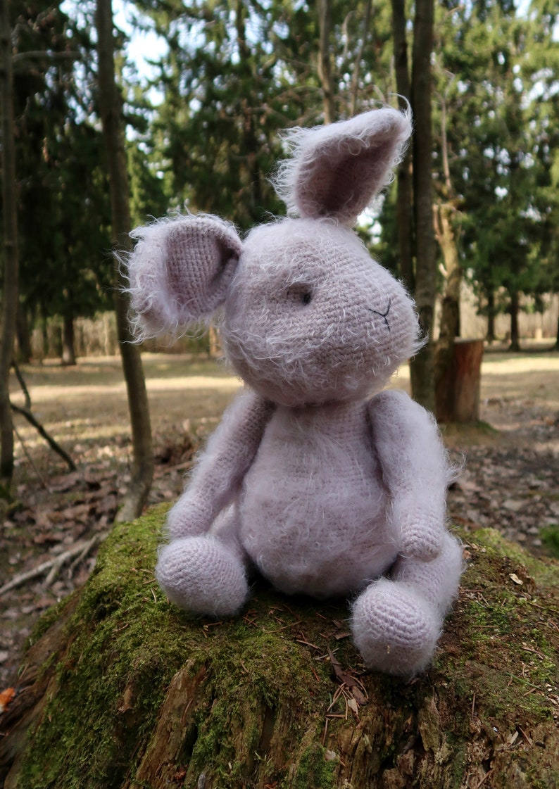 Op de afbeelding: Een zacht, pluche, lichtpaarse gehaakte konijnenknuffel die op een mossige boomstronk in een bosrijke omgeving zit.
