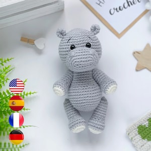 Op de afbeelding: Een grijs gehaakt nijlpaard speelgoed met witte accenten op de voeten en oren. Het speelgoed ligt op een wit oppervlak met een groene plant, een houten ster en een wit gehaakt deken op de achtergrond.