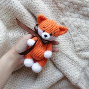 Puede incluir: Un peque&ntilde;o juguete de zorro tejido a ganchillo hecho a mano con hilo naranja brillante, detalles blancos en las patas y el hocico, ojos de cuentas negras y una cinta de sat&eacute;n marr&oacute;n oscuro atada al cuello. El texto @KNIT.FRIENDS es visible.