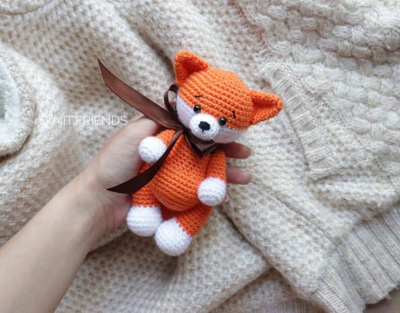 Puede incluir: Un peque&ntilde;o juguete de zorro tejido a ganchillo hecho a mano con hilo naranja brillante, detalles blancos en las patas y el hocico, ojos de cuentas negras y una cinta de sat&eacute;n marr&oacute;n oscuro atada al cuello. El texto @KNIT.FRIENDS es visible.