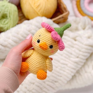 May include: A small, yellow crocheted duckling toy featuring a pink flower-shaped hat with a green stem top. The duckling has black bead eyes, a small orange beak, and orange feet. The body is crafted from yellow yarn with visible stitches.