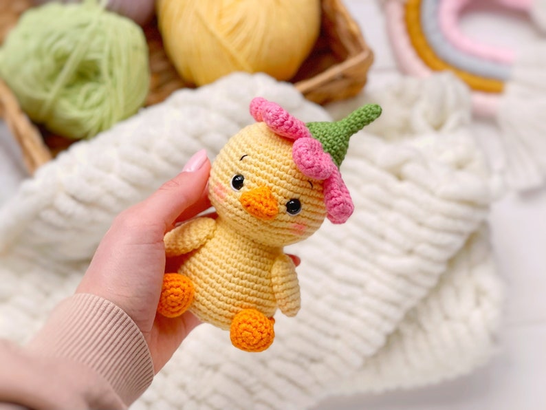 May include: A small, yellow crocheted duckling toy featuring a pink flower-shaped hat with a green stem top. The duckling has black bead eyes, a small orange beak, and orange feet. The body is crafted from yellow yarn with visible stitches.