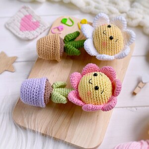 May include: Two crocheted flower pot toys with smiling faces. One flower is white with a yellow center and the other is pink with a yellow center. Both are in brown pots with green stems.