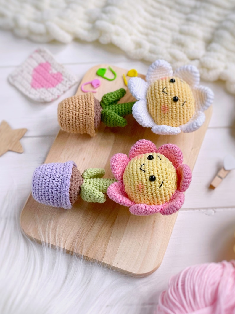 May include: Two crocheted flower pot toys with smiling faces. One flower is white with a yellow center and the other is pink with a yellow center. Both are in brown pots with green stems.