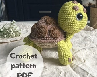 Puede incluir: Patrón de crochet PDF para un juguete de tortuga de crochet verde y marrón. La tortuga tiene una cabeza grande con un ojo grande y un caparazón marrón.