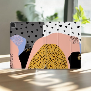 Puede incluir: Una funda para iPad con un diseño abstracto colorido. La funda presenta un patrón de puntos negros sobre un fondo gris, con formas rosas, amarillas y azules. La funda está abierta, revelando la pantalla del iPad.