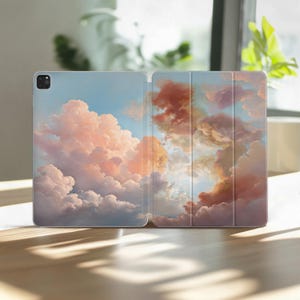 Könnte beinhalten: Geöffnete Tablet-Hülle mit Wolkenlandschaft. Die Hülle zeigt einen pastellfarbenen Himmel mit flauschigen rosa, orangefarbenen und weißen Wolken vor blauem Hintergrund. Die Hülle ist geöffnet und zeigt das Innere und den Bildschirm.