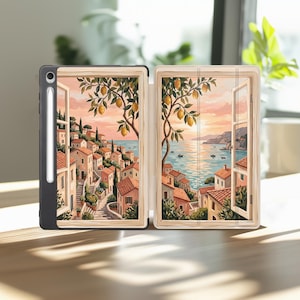 May include: A black tablet case with a stylus holder featuring a split-panel illustration of a coastal village at sunset. The artwork depicts terracotta-roofed buildings, stone staircases, lemon trees, and a calm sea viewed through open windows.