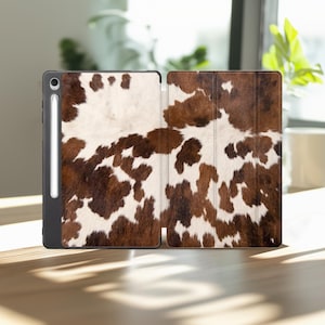 May include: An open tablet case with a cowhide pattern. The case is brown and white, with a black border. A stylus is attached to the side of the tablet. The case is open, revealing the interior.