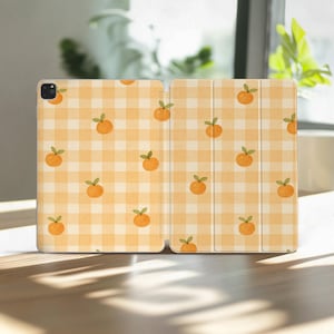 May include: An orange and white plaid iPad case with a repeating pattern of orange fruit with green leaves. The case is open, revealing the interior and exterior design. The case is on a wooden surface.