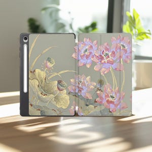 May include: A tablet case featuring a sage green background with a botanical print of pink lotus flowers and gold-toned leaves. The case includes a dedicated slot for a white stylus pen, showcasing a tri-fold design that covers the front and back.