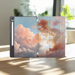 May include: A black tablet case with a pastel cloud design. The case is open, revealing the tablet screen and a stylus. The cloud design features shades of pink, orange, and blue, creating a dreamy aesthetic. The case is on a wooden surface.