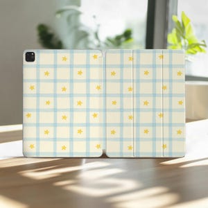 May include: An open tablet case with a light beige background, featuring a blue plaid pattern and scattered yellow stars. The case is designed to protect the tablet and has a magnetic closure.