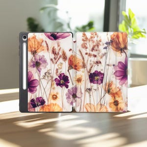 May include: A tablet case featuring a floral design with purple, orange, and yellow flowers. The case is open, revealing the interior with a stylus attached. The background is white, and the case is on a wooden surface.