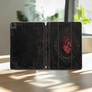 May include: Black iPad case with an open book design. The left side is plain with ornate corner designs. The right side features a red anatomical heart within an oval frame, surrounded by decorative scrollwork.