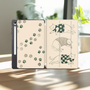 May include: A tablet case with a cream-colored exterior featuring a pattern of dark green and light gray paw prints on the left side. The right side displays three illustrations of cats wearing patterned shirts, including floral, striped, and checkered designs.