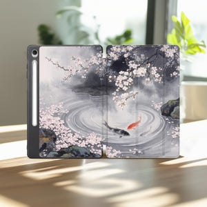 Può includere: Una custodia per tablet con un'illustrazione stampata di uno stagno con pesci koi e rami di ciliegio. Il design include un pesce koi nero e uno rosso che nuotano in acqua con increspature, circondati da petali di fiori rosa e rocce scure.
