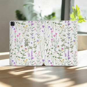 Puede incluir: Una funda blanca para tableta con un colorido estampado floral. El diseño incluye varias flores silvestres en tonos rosa, morado, amarillo y verde. La funda está abierta, mostrando el diseño interior, y se encuentra sobre una superficie de madera.