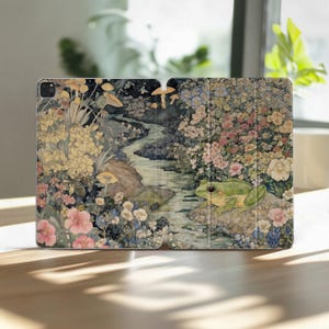 Könnte beinhalten: Eine iPad-Hülle mit einer skurrilen Illustration einer ruhigen Landschaft mit einem Bach, Blumen und einem grünen Frosch. Das Kunstwerk hat eine Vintage-Ästhetik mit sanften Farben und Pilzdetails.