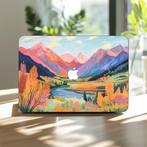 May include: A laptop case displaying a scenic mountain landscape. The design showcases a mountain range in shades of pink, blue, and orange, a river, and trees. The Apple logo is visible on the case.