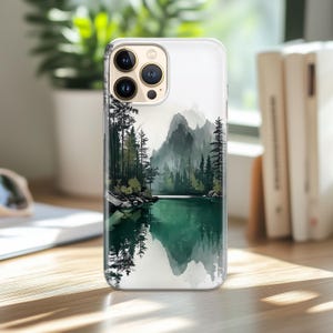 May include: A clear phone case featuring a watercolor landscape of a lake surrounded by evergreen trees and mountains. The water reflects the trees and mountains. The case has a camera cutout and buttons.