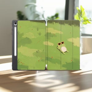 May include: A black tablet case with a green illustrated cover featuring a small frog wearing red sunglasses lying in a grassy field. The case includes a dedicated slot for a white stylus pen on the left side, maintaining a sleek profile.
