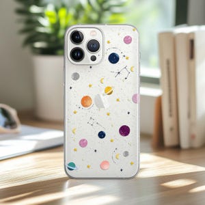 May include: Clear phone case with a space theme. The case has colourful planets, stars, and constellations on a white background. The planets are in shades of orange, blue, purple, and pink. The case is on a wooden surface.