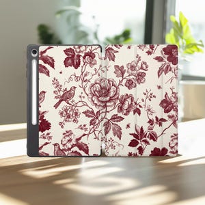 May include: A tablet case with a cream-colored background featuring a dark red botanical print of flowers, leaves, and birds. The case includes a dedicated slot for a stylus on the left side, and the cover displays a continuous floral pattern.