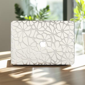Puede incluir: Un portátil blanco con un diseño floral. La cubierta presenta un patrón repetido de flores tipo margarita en un diseño texturizado en relieve. El logotipo de Apple es visible. El portátil está sobre una superficie de madera.