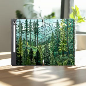 May include: A tablet case featuring a stained glass style design of a dense forest with tall pine trees and a mountain range in the background. The case is black with a white stylus pen attached to the side, displaying a vibrant green color palette.